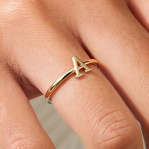 Op de afbeelding: Een dunne, gepolijste goudkleurige ring met een opvallende hoofdletter A in het midden van de band. De ring heeft een gladde, reflecterende metalen afwerking en een eenvoudig, slank ontwerp dat horizontaal op de vinger rust.