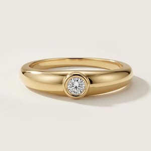 May include: A polished gold ring featuring a smooth, rounded band with a single, round, brilliant-cut clear gemstone set securely in a bezel setting at the centre. The metal has a high-shine finish, reflecting light across its curved surface.