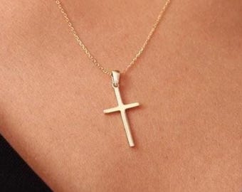 May include: A gold-toned metal cross pendant suspended from a delicate, matching gold-toned chain. The cross features a simple, smooth, and polished design with clean lines, resting against a dark fabric background.