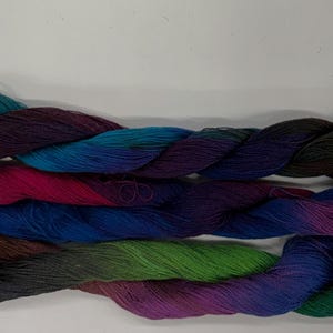 Può includere: Tre matasse di filato tinto a mano disposte orizzontalmente. La matassa superiore sfuma dal marrone scuro al verde acqua e viola intenso. Quella centrale presenta magenta, blu reale e viola scuro. Quella inferiore mescola marrone, verde e viola.
