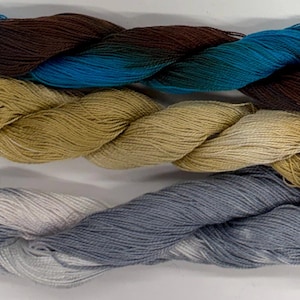 Può includere: Tre matasse di filato disposte orizzontalmente. La matassa superiore presenta una sfumatura di blu scuro, marrone e verde acqua. La matassa centrale è di colore beige tinta unita con un'etichetta bianca con il numero 26. Quella inferiore è grigia.