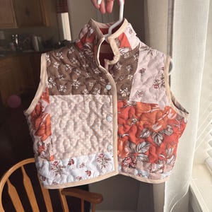 May include: A sleeveless quilted vest featuring a patchwork design with various floral and polka dot patterns in shades of burnt orange, brown, cream, and white. The vest has a high collar, light beige trim, and three white snap button closures.