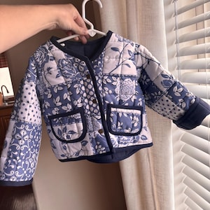 May include: A quilted jacket featuring a blue and white patchwork pattern with floral and polka dot motifs. The jacket has a round neckline, long sleeves, two front patch pockets, and a dark blue trim along the edges and button closure.