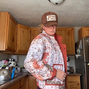 May include: A brown trucker hat with a patch featuring a horse graphic and the text "SADDLE UP" and a quilted jacket with a patchwork floral pattern in shades of orange, white, and brown, featuring a front button closure and a patch pocket.