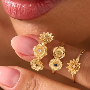Puede incluir: Primer plano de varios anillos florales dorados. Cada anillo presenta un diseño floral diferente, algunos con pequeñas piedras preciosas. Los anillos se llevan en los dedos, mostrando los intrincados detalles y la artesanía de la joyería.