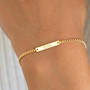 May include: A gold-colored bracelet with a delicate chain and a rectangular bar. The bar is engraved with the initials "GOC" and a small heart symbol. The bracelet is worn on a wrist.