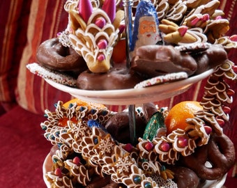 May include: A three-tiered serving stand filled with gingerbread cookies, chocolate-covered pretzels, and mandarin oranges. The gingerbread cookies are decorated with white icing and red and green sprinkles. The chocolate-covered pretzels are arranged in a spiral pattern. The mandarin oranges are arranged in a circle around the base of the stand.