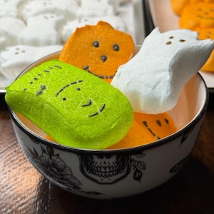 May include: A white ceramic bowl decorated with black floral and skull illustrations contains three marshmallow candies. The candies include a bright green rectangular shape, a white ghost shape, and an orange pumpkin shape, each with black facial features.