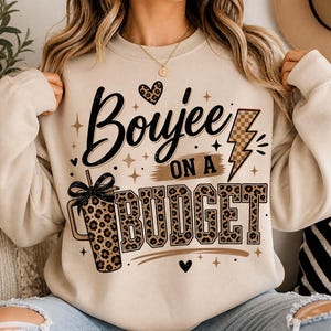 May include: Cream-colored sweatshirt with the phrase "Boujee on a Budget" in black script and leopard print. The design includes a leopard print cup, a lightning bolt, and heart accents. The sweatshirt is a casual, comfortable style.