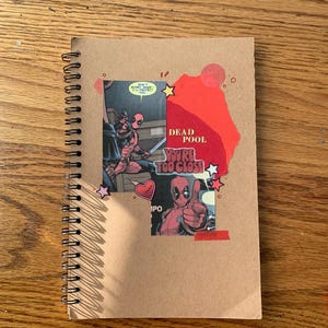 Puede incluir: Una libreta de espiral marrón con un collage de imágenes de cómics de Deadpool. El collage incluye un corazón rojo, estrellas y el texto "Dead Pool You're Too Close".