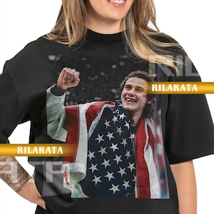 May include: Black adult unisex t-shirt featuring a graphic of a person wrapped in an American flag. The shirt has the text "RILARATA" in gold lettering. The shirt is a ready custom design.