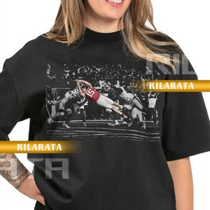 Puede incluir: Camiseta unisex negra para adultos con una imagen en escala de grises de un jugador de fútbol americano en acción. El jugador lleva un uniforme rojo y blanco. La camiseta tiene el texto "RILARATA" y "READY CUSTOM DESIGNS".