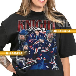 Könnte beinhalten: Schwarzes Unisex-T-Shirt für Erwachsene mit einer Grafik eines Hockeyteams, dem Text "KNIGHT Hilary" in Rot und Weiß und dem USA-Logo. Das Team trägt blaue und weiße Uniformen. Das Shirt hat auch den Text "RILARATA".