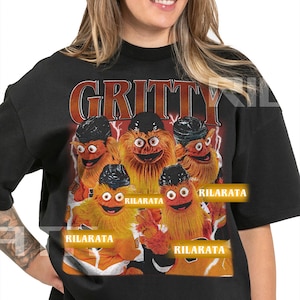 Op de afbeelding: Zwart unisex T-shirt voor volwassenen met een afbeelding van de mascotte Gritty en het woord "GRITTY" boven de afbeelding. De afbeelding bevat de tekst "RILARATA" op het shirt. Het shirt is een kant-en-klaar custom design.