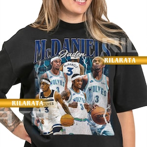 Op de afbeelding: Zwart unisex T-shirt voor volwassenen met een afbeelding van basketballer Jaden McDaniels in een Wolves-shirt. Het ontwerp bevat de tekst "McDaniels" en "Wolves" met nummer 3. Het shirt heeft ook de tekst "Rilarata" en "Ready Custom Designs".