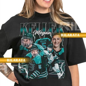 Könnte beinhalten: Schwarzes Unisex-T-Shirt für Erwachsene mit einem teal- und weißen Hockey-Design. Das Shirt zeigt den Namen "Keller" und "Megan" sowie den Text "Rilarata" und "Ready Custom Designs".