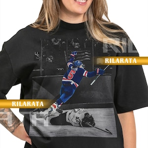 May include: Black adult unisex t-shirt featuring a hockey action scene. The graphic shows a player in a blue and white uniform with the number 5, and another player on the ice. Text includes "RILARATA" and "READY CUSTOM DESIGNS".