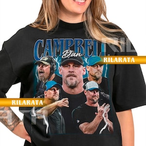 May include: Black unisex t-shirt featuring a graphic of a man with the text "CAMPBELL Dan" in blue. The design includes multiple images of the man. The shirt has the text "RILARATA" and "ADULT UNISEX TSHIRT" printed on it.