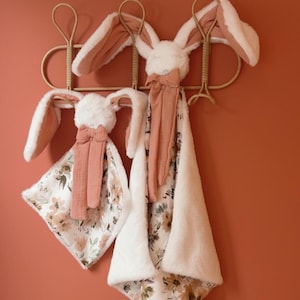 May include: Two plush rabbit security blankets featuring white faux fur heads with long ears, attached to soft fabric squares. One side of the blankets displays a floral pattern in muted earth tones, while the other side is solid white faux fur.