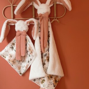 May include: Two plush rabbit security blankets featuring soft white faux fur heads with long ears, accented by terracotta-colored fabric bows. The blankets have a floral print on one side and a solid white plush texture on the reverse side.