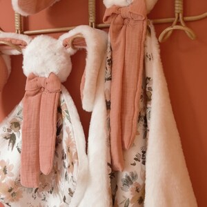 May include: Two plush security blankets featuring rabbit heads with long, soft white faux fur ears and terracotta-colored fabric accents. The blankets have a floral pattern on one side and a solid white faux fur texture on the reverse side.