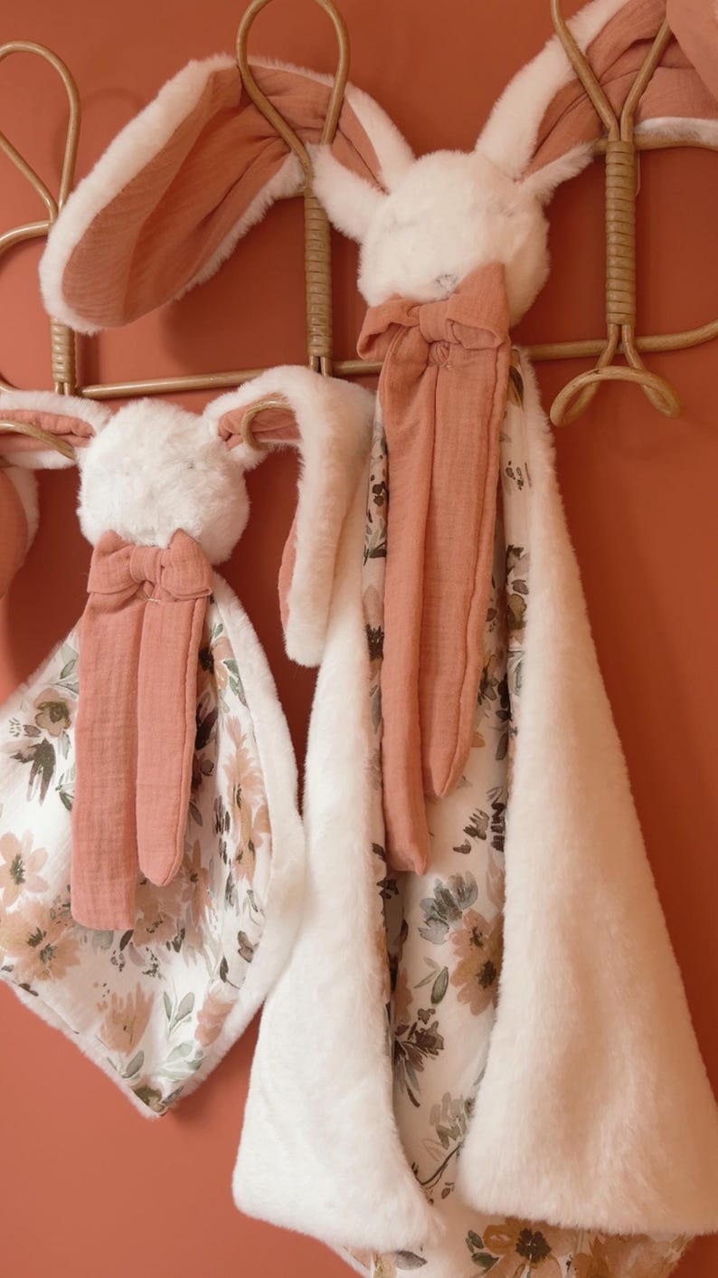 May include: Two plush security blankets featuring rabbit heads with long, soft white faux fur ears and terracotta-colored fabric accents. The blankets have a floral pattern on one side and a solid white faux fur texture on the reverse side.