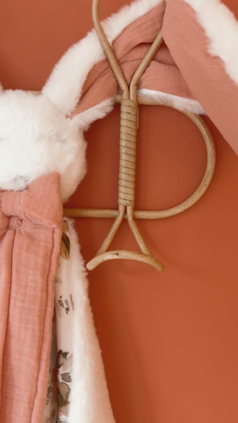 May include: A soft, plush security blanket featuring a white faux fur texture and a dusty rose-colored fabric with a floral pattern. The blanket is draped over a light brown, curved rattan wall hook, showcasing its layered, multi-textured design.