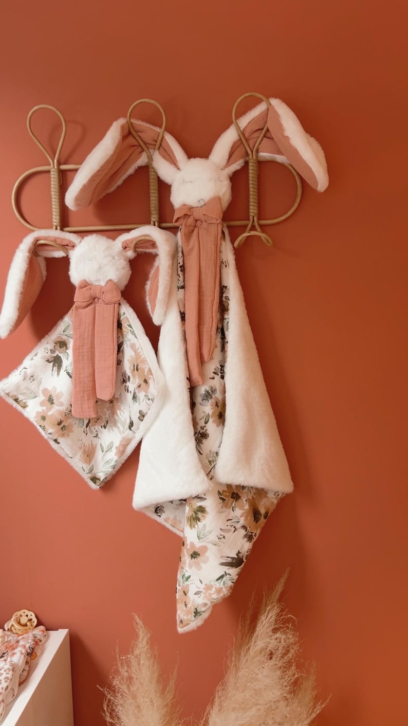 May include: Two plush rabbit security blankets featuring soft white faux fur heads with long ears, accented by terracotta-colored fabric bows. The blankets have a floral print on one side and a solid white plush texture on the reverse side.