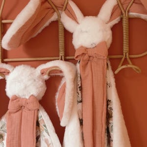 May include: Two plush rabbit security blankets featuring soft white faux fur heads with long ears lined in terracotta fabric. Each blanket has a matching terracotta bow at the neck and a fabric body with a floral pattern in shades of brown and green.