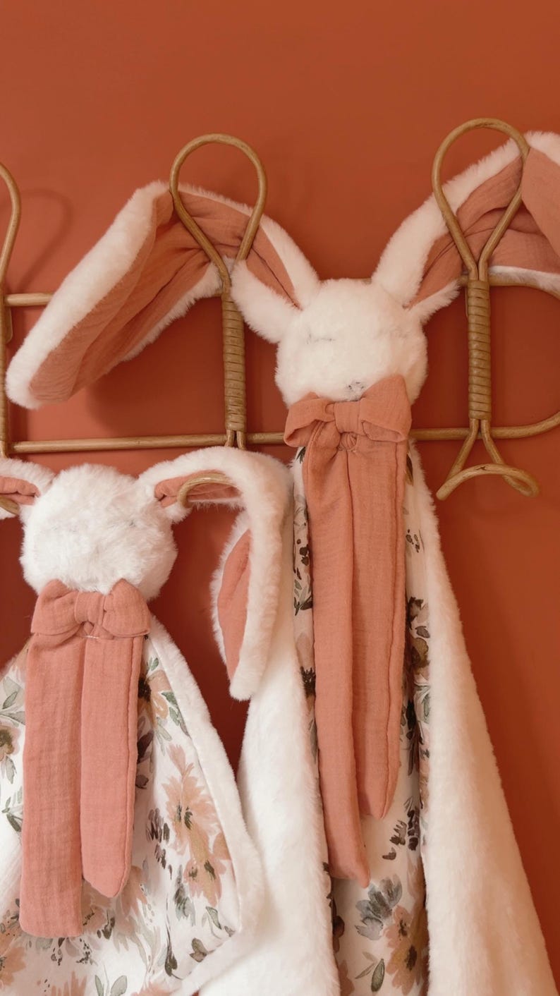May include: Two plush rabbit security blankets featuring soft white faux fur heads with long ears lined in terracotta fabric. Each blanket has a matching terracotta bow at the neck and a fabric body with a floral pattern in shades of brown and green.