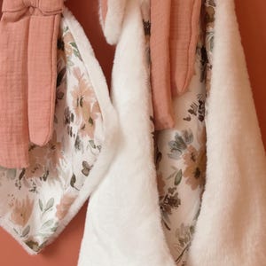 May include: A plush white rabbit security blanket featuring long ears and a bow, paired with a matching rectangular blanket. Both items are crafted from a combination of soft white faux fur and a white fabric printed with a floral pattern in muted tones of dusty rose, sage green, and brown.