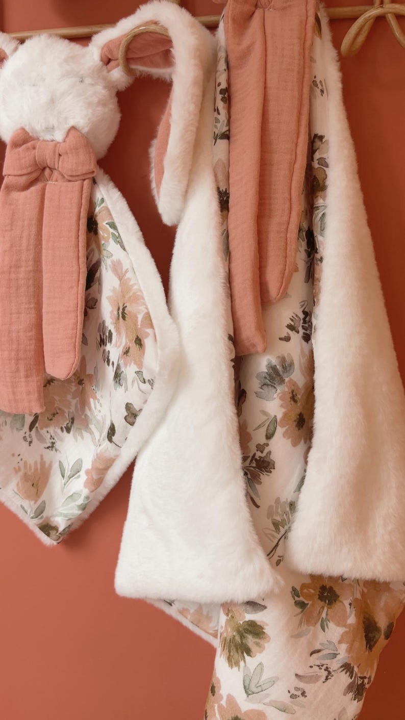 May include: A plush white rabbit security blanket featuring long ears and a bow, paired with a matching rectangular blanket. Both items are crafted from a combination of soft white faux fur and a white fabric printed with a floral pattern in muted tones of dusty rose, sage green, and brown.