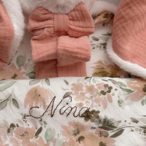 May include: A plush white rabbit toy with long ears featuring a soft peach-colored fabric lining and a matching peach bow. The rabbit is paired with a white blanket decorated with a floral pattern in shades of peach and green, embroidered with the name Nina.