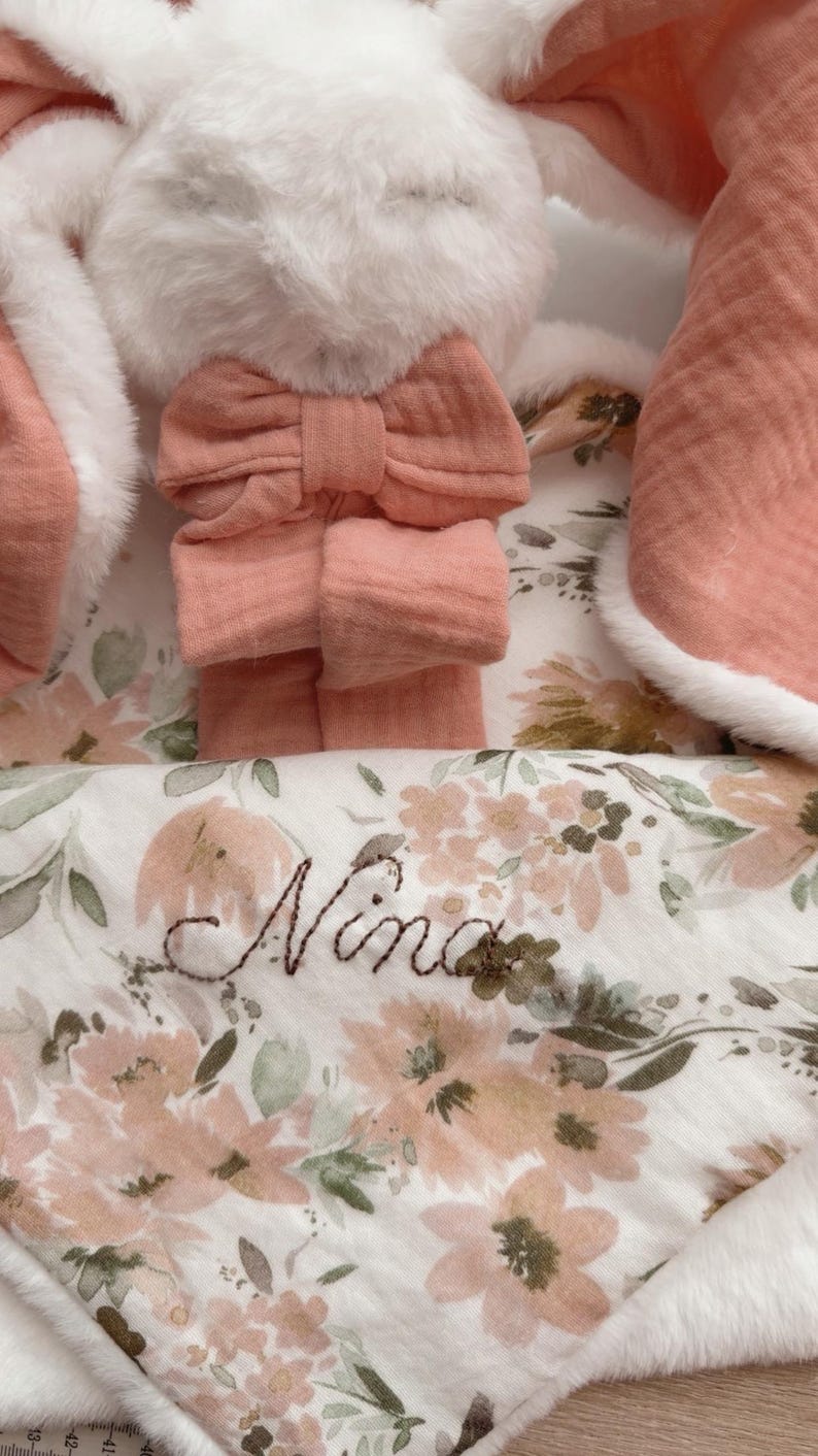 May include: A plush white rabbit toy with long ears featuring a soft peach-colored fabric lining and a matching peach bow. The rabbit is paired with a white blanket decorated with a floral pattern in shades of peach and green, embroidered with the name Nina.
