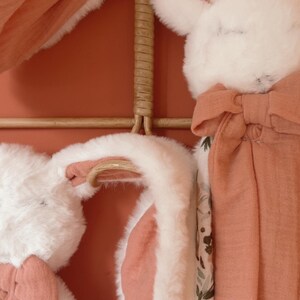 May include: Two plush rabbit security blankets featuring soft white faux fur heads with long ears, attached to terracotta-colored muslin fabric squares. One side of the fabric features a floral pattern with muted green and brown tones.