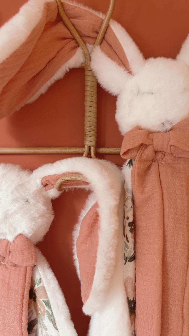 May include: Two plush rabbit security blankets featuring soft white faux fur heads with long ears, attached to terracotta-colored muslin fabric squares. One side of the fabric features a floral pattern with muted green and brown tones.