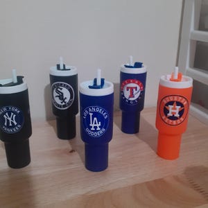 May include: A collection of five insulated tumblers featuring logos of various Major League Baseball teams. The tumblers are in different colors, including black, blue, and orange, with white lids and straws. Each tumbler displays a team logo, such as the Yankees or Dodgers.