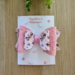 May include: A hair bow featuring layers of pink glitter fabric and a white patterned material with cartoon characters and food items. The center is cinched with a pink ribbon. The bow is attached to a white display card labeled Angelina's Bowtique.