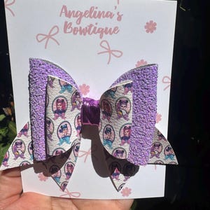 May include: A hair bow featuring a layered design with textured purple glitter fabric and a white faux leather layer printed with a repeating pattern of cartoon characters in oval frames. A metallic purple center band holds the layers together.