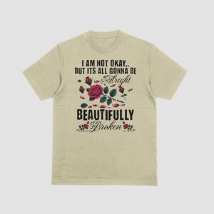May include: Beige t-shirt with the text "I AM NOT OKAY.. BUT ITS ALL GONNA BE Alright BEAUTIFULLY Broken" in black and a red rose graphic. The graphic includes red roses and green leaves.