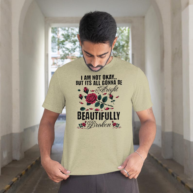 May include: A light tan t-shirt with the text "I AM NOT OKAY.. BUT ITS ALL GONNA BE Alright BEAUTIFULLY Broken" in black and red. The design includes a rose and leaves.
