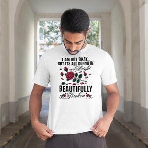 May include: A light gray t-shirt with the text "I AM NOT OKAY.. BUT ITS ALL GONNA BE Alright BEAUTIFULLY Broken" in black and red. The design includes a red rose and rosebuds.