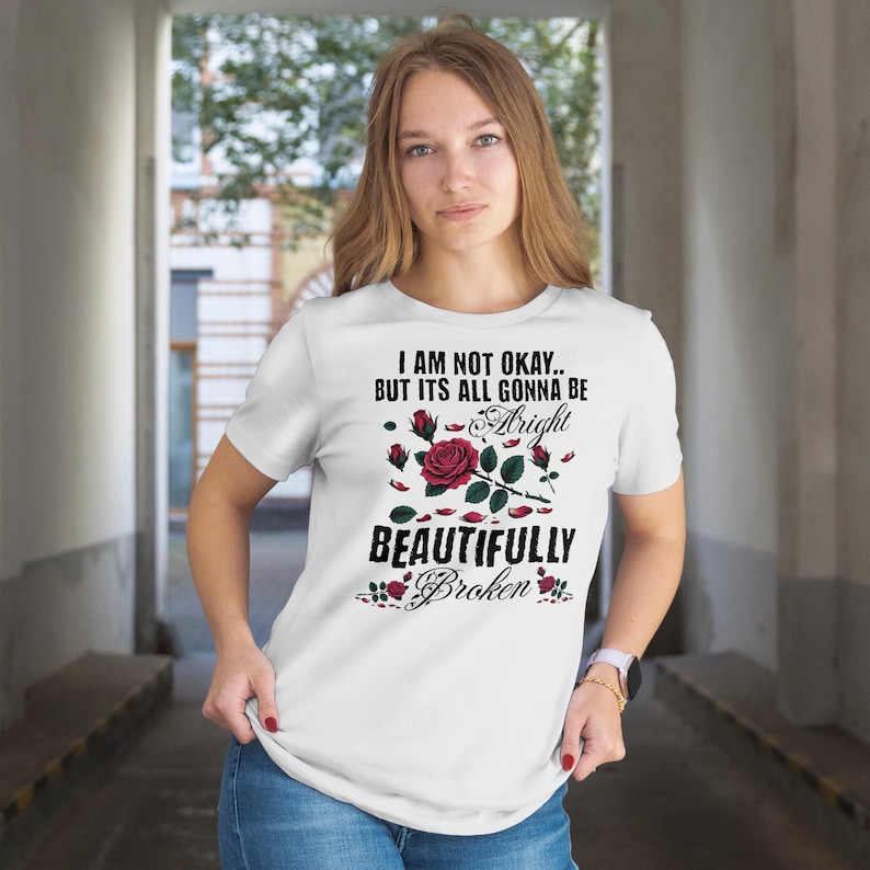 May include: White t-shirt with the text "I AM NOT OKAY.. BUT ITS ALL GONNA BE Alright BEAUTIFULLY Broken" in black and a floral design with red roses and green leaves. The shirt is short-sleeved.