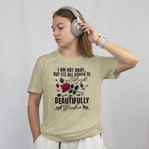 May include: A light beige t-shirt with the text "I AM NOT OKAY.. BUT ITS ALL GONNA BE Alright BEAUTIFULLY Broken" in black script and a floral design with red roses. The shirt is short-sleeved.