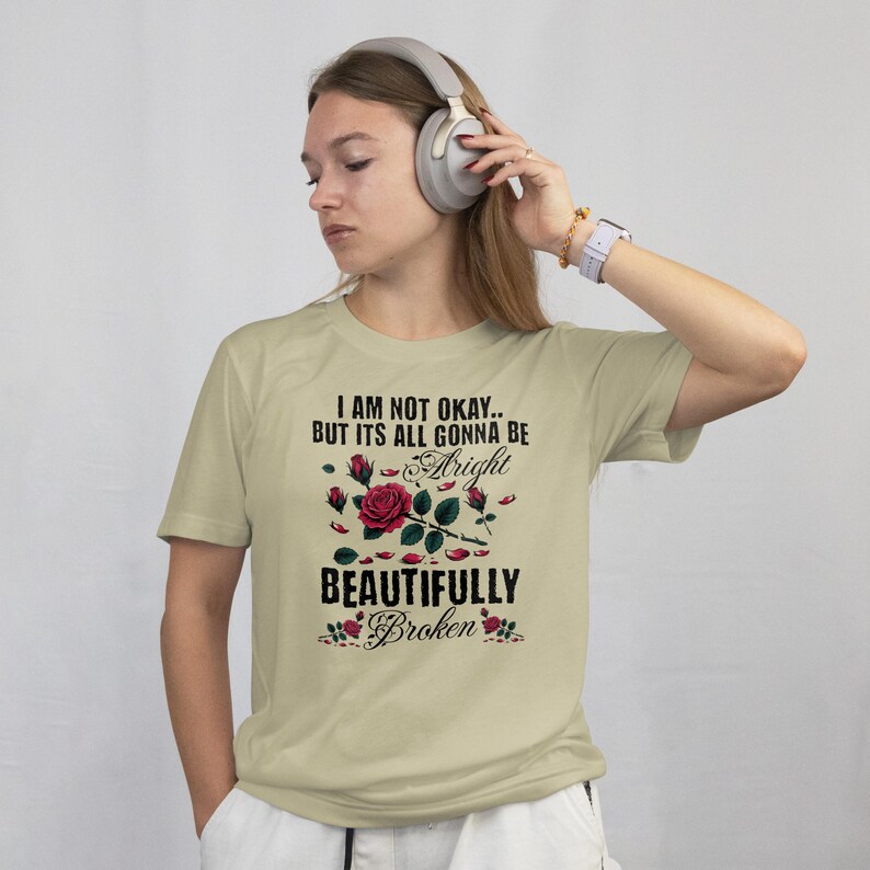 May include: A light beige t-shirt with the text "I AM NOT OKAY.. BUT ITS ALL GONNA BE Alright BEAUTIFULLY Broken" in black script and a floral design with red roses. The shirt is short-sleeved.