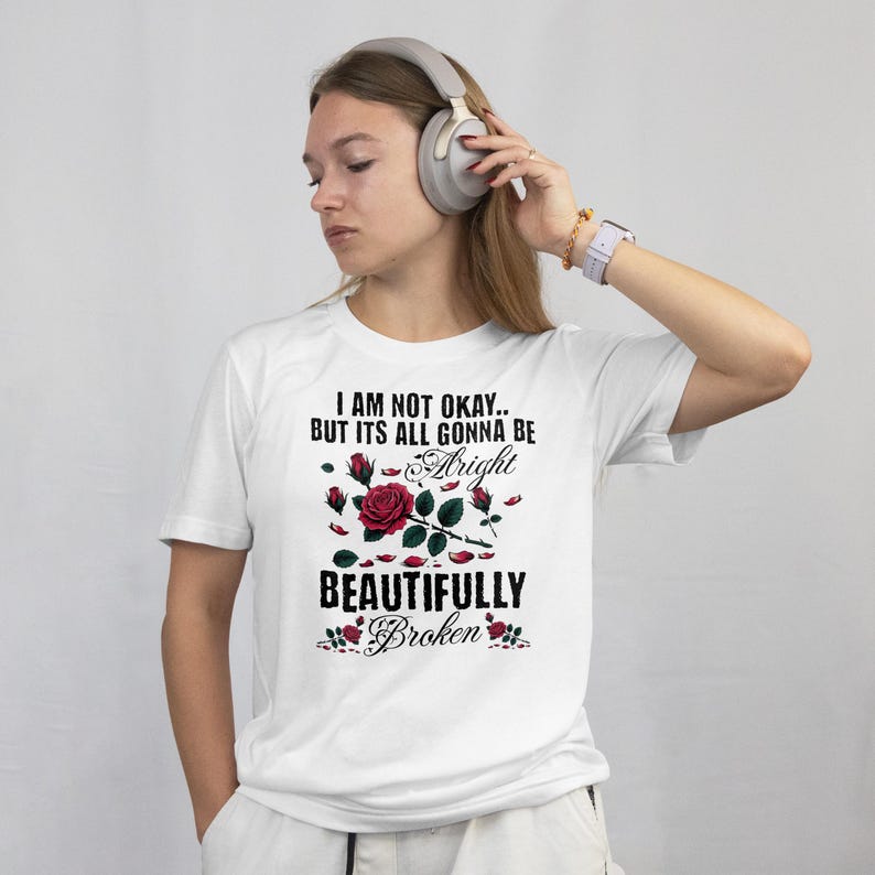 May include: White t-shirt with the text "I AM NOT OKAY.. BUT ITS ALL GONNA BE Alright BEAUTIFULLY Broken" and a rose graphic. The shirt is being worn by a person wearing headphones and a watch.
