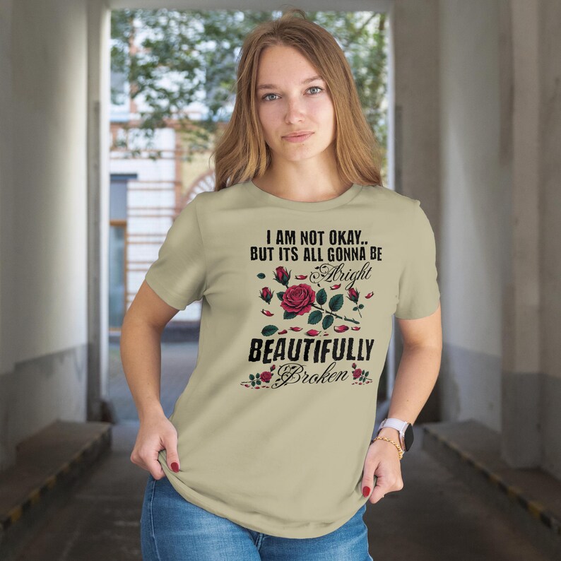 May include: A beige t-shirt with the text "I AM NOT OKAY.. BUT ITS ALL GONNA BE Alright BEAUTIFULLY Broken" and a rose graphic. The graphic includes a red rose, rose petals, and smaller red roses. The shirt is short-sleeved.