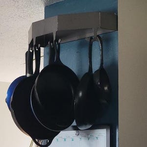 May include: A set of five black cast iron skillets hanging from a metal rack mounted on a wall. The skillets are of varying sizes and are all hanging upside down.