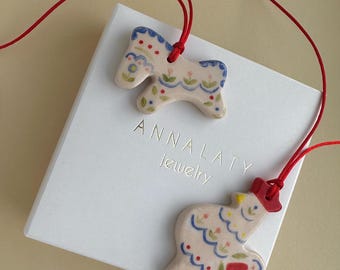 May include: Two ceramic pendants on red cords, one shaped like a horse and the other like a rooster, both with colorful floral patterns. The pendants are displayed on a white box with the text "ANNALATY jewelry".