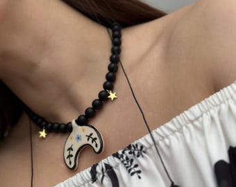 May include: A black beaded necklace with a ceramic crescent moon pendant. The pendant is decorated with floral designs and a blue eye. The necklace has gold star charms.